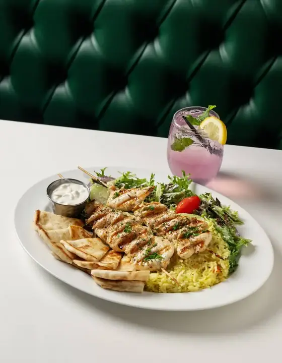 Grilled chicken souvlaki skewers with pita, rice, salad, and tzatziki beside a lavender lemonade