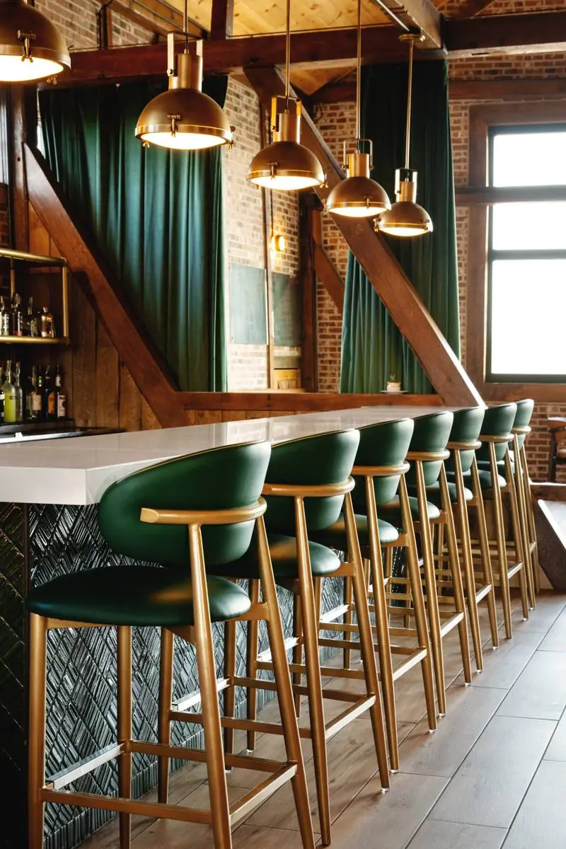 The bar with green leather and brass stools beneath brass pendant lights, with green velvet curtains and exposed brick behind